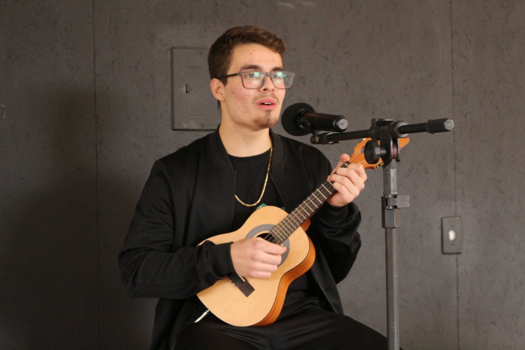 Homem com violão canta música sentado em frente ao microfone 